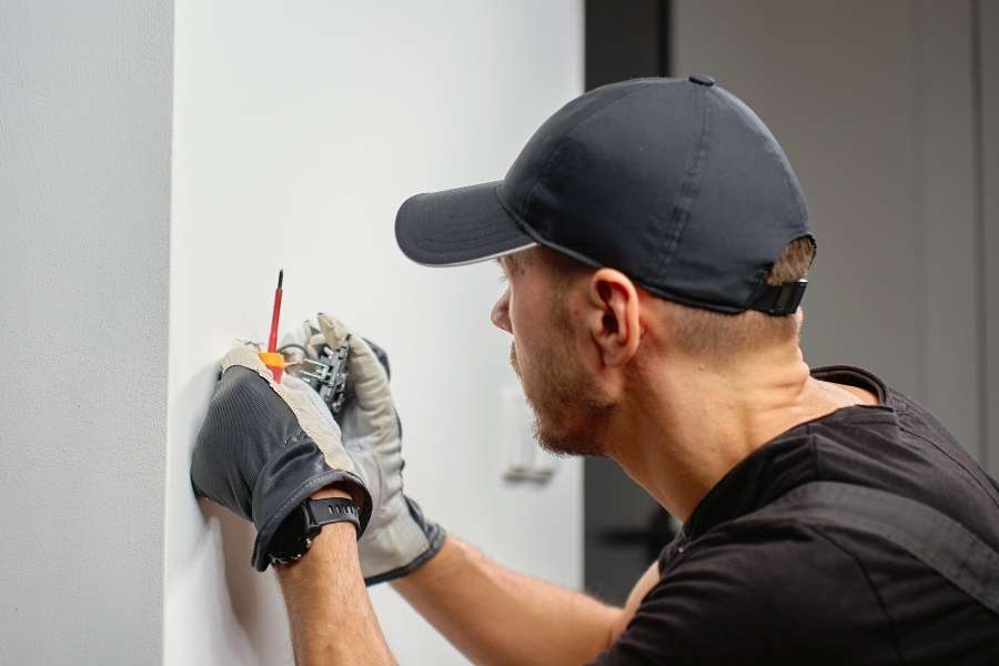 Un technicien portant des gants de protection et une casquette noire installe un petit mécanisme électrique ou un interrupteur dans un boîtier encastré sur un mur blanc.